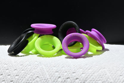 A group of silicone scissor finger ring in neon green, purple, and black, arranged on a white textured surface against a black background. The purple plugs show the brand name 'RW PROGRESSIVE GEAR' embossed on them.